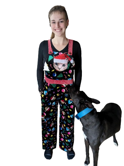 Molly, wearing a pair of overalls printed with cats in Santa hats, ornaments, stars, Christmas trees, and candy. There is a large, slightly puffy applique of the cat with the Santa hat on the bib, and the straps are red with white polka dots. Atlas, a large mostly black pit bull/husky/german shepherd mix, is standing next to Molly, looking up at the cat on the bib with some alarm.