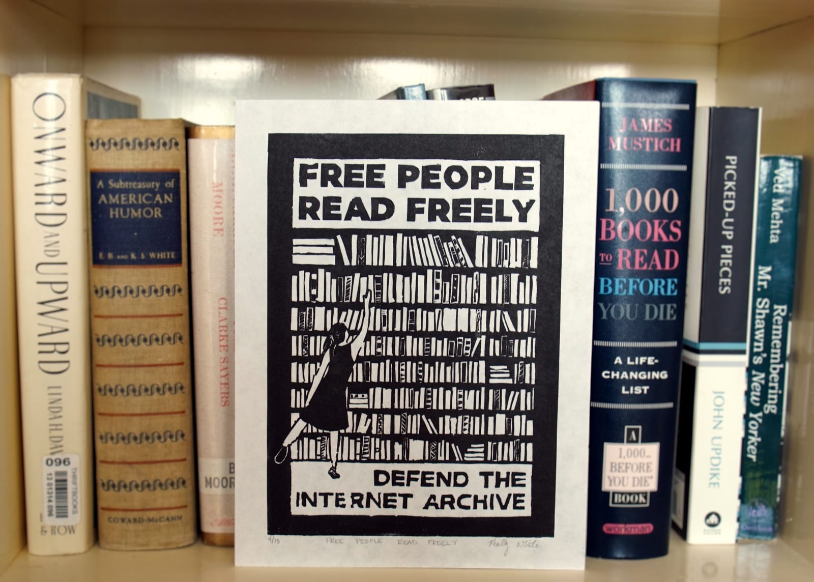 A 6x8" linocut print in black ink depicting a person in a dress reaching for a book on a large bookshelf. Above is “Free people read freely”; below is “Defend the Internet Archive”. The print is propped up against a shelf of books.